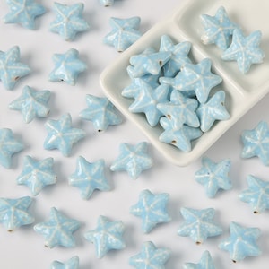 May include: A collection of light blue ceramic starfish beads. The beads are scattered on a white surface and in a white divided dish.