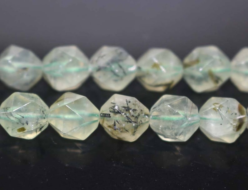 May include: A strand of faceted green and black gemstone beads. The beads are faceted and have a starburst shape. The beads are strung on a clear elastic cord.