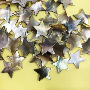 Puede incluir: Un montón de abalorios de concha de estrella de color marrón iridiscente con dos agujeros en cada estrella. Los abalorios están esparcidos sobre una superficie amarilla.