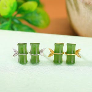 May include: Green jade earrings shaped like bamboo stalks. The earrings have silver or gold-coloured bands and leaf details. The bamboo design is a vibrant green colour, with a polished finish.