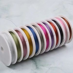 Puede incluir: Un conjunto de 10 carretes de alambre de colores, cada uno enrollado alrededor de un carrete de plástico blanco. Los colores incluyen plata, verde, rosa, oro, azul, gris, rosa, morado, cobre y negro.