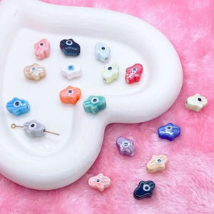 May include: Assortment of colorful hamsa hand charms with an evil eye design. The charms are made of glass and have a variety of colors, including blue, green, pink, purple, and white.