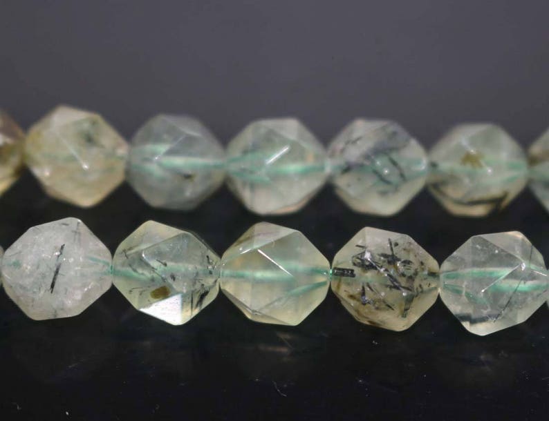 May include: A strand of faceted green and clear quartz beads with black inclusions. The beads are strung on a green thread.