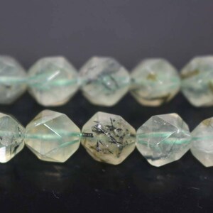 May include: A strand of faceted green and black gemstone beads. The beads are faceted and have a starburst shape. The beads are strung on a clear elastic cord.