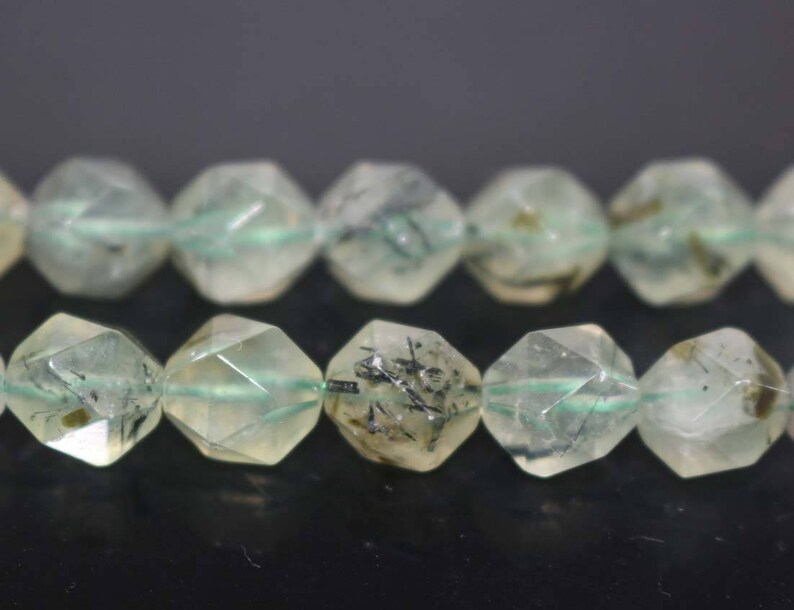 May include: A strand of faceted green and black gemstone beads. The beads are faceted and have a starburst shape. The beads are strung on a clear elastic cord.
