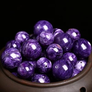 May include: A close-up of a collection of purple and white gemstone beads. The beads are round and have a smooth, polished finish. They are arranged in a bowl.