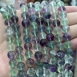 May include: Strands of round, polished fluorite beads in shades of green, purple, and clear. The beads are strung together, showcasing the natural color variations and patterns within the stone. The beads are approximately 8mm in diameter.