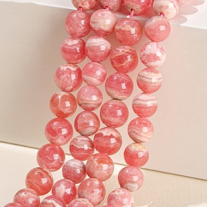 Puede incluir: Una hebra de cuentas redondas de color rosa y blanco con rayas. Las cuentas están hechas de piedra natural y tienen un acabado pulido.