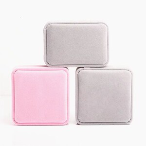 May include: Three small, square jewelry boxes in a stack. One box is light pink, and the other two are gray. The boxes are made of a soft, velvety material and have rounded corners. The boxes are empty and ready to be filled.
