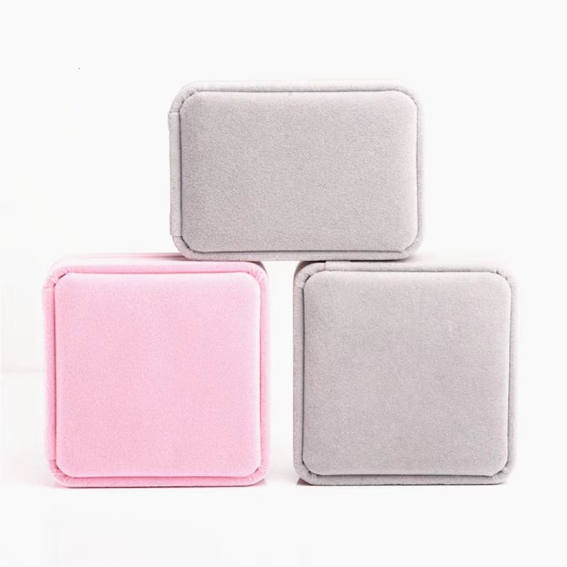 May include: Three small, square jewelry boxes in a stack. One box is light pink, and the other two are gray. The boxes are made of a soft, velvety material and have rounded corners. The boxes are empty and ready to be filled.