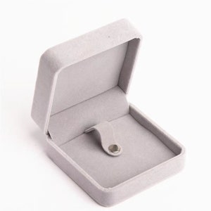 May include: Open, light gray jewelry box with a soft, velvety texture. The box is rectangular with a hinged lid and a small, curved holder inside. A silver-colored snap secures the lid.