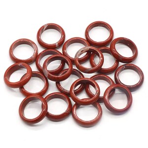 May include: A pile of 15 red jasper stone rings. The rings are smooth and polished, with a natural red and white marbled pattern.