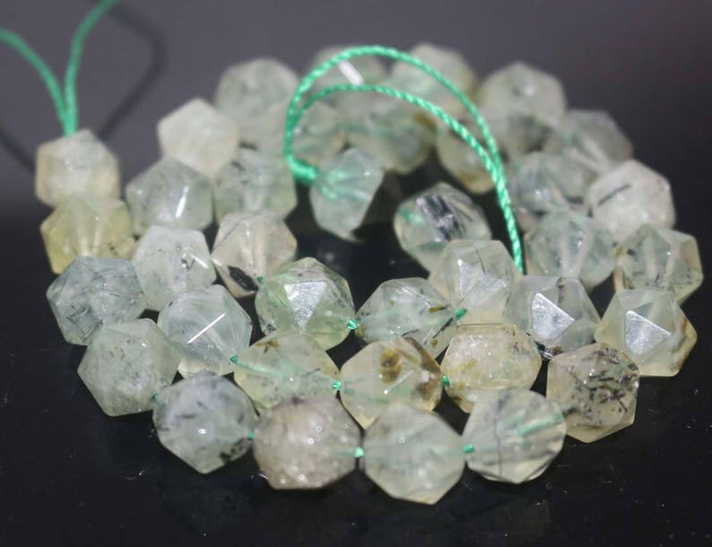 May include: A strand of green and black gemstone beads. The beads are faceted and have a translucent appearance. The beads are strung on a green thread.