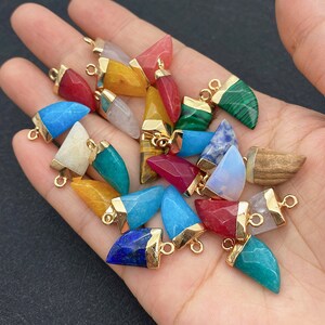 May include: A collection of gemstone pendants in various colors, including red, blue, yellow, and green. Each pendant has a gold-colored cap and a loop for attaching to a chain or other jewelry component. The gemstones have a polished finish.