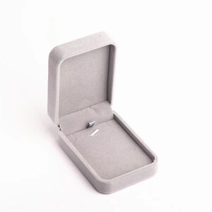 May include: An open, gray velvet jewelry box. The box is rectangular with rounded corners and a hinged lid. The interior is lined with matching gray velvet, and a small metal clasp secures the lid. The box is designed to hold a necklace or pendant.