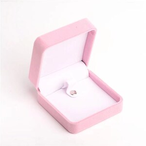 May include: A light pink velvet jewelry box, open to reveal a white interior. The box has a square shape with a hinged lid and a small, silver-colored clasp. The interior features a white, padded lining.