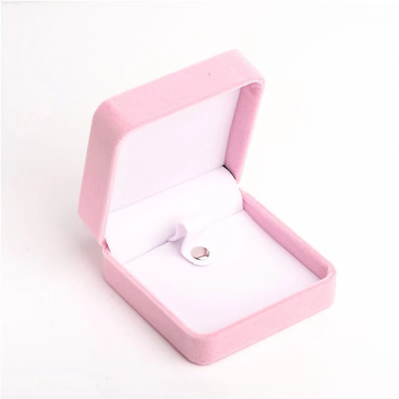 May include: A light pink velvet jewelry box, open to reveal a white interior. The box has a square shape with a hinged lid and a small, silver-colored clasp. The interior features a white, padded lining.