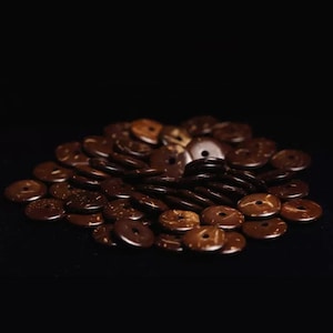May include: A pile of brown coconut shell buttons. The buttons are round and have a hole in the centre. They are arranged in a random pattern.