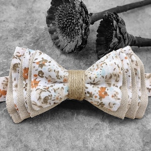Può includere: Un papillon bianco e marrone con un motivo floreale. Il papillon è realizzato in tessuto e ha un aspetto rustico e fatto a mano.