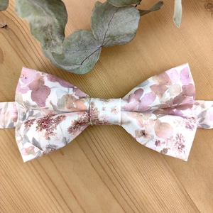 Könnte beinhalten: Eine Fliegen aus Stoff mit floralem Muster in Creme, Rosa, Braun und Lila. Die Fliege wird auf einer Holzoberfläche präsentiert. Im Hintergrund ein Eukalyptuszweig. Geeignet für formelle Anlässe.