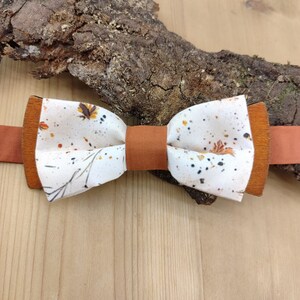 Könnte beinhalten: Eine handgefertigte Fliege mit einem weißen Stoffmittelteil mit einem floralen Muster in Braun und Orange. Die Fliege hat braune, lederartige Enden und ein passendes braunes Mittelband. Der Hintergrund ist eine Holzoberfläche mit einem Stück Rinde.