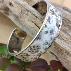 Op de afbeelding: Een zilverkleurige manchetarmband met een bloemenstoffen inleg. De stof heeft een witte achtergrond met blauwe, bruine en groene bloempatronen. De armband wordt gepresenteerd op een stuk drijfhout, met donkerrood en groen blad op de voorgrond.