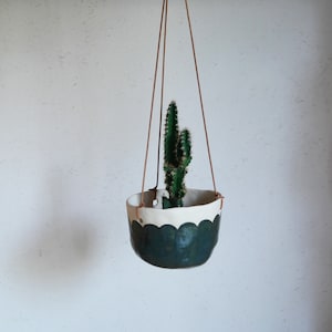 Peut inclure: Un pot suspendu en céramique vert et blanc avec un bord festonné, contenant un petit cactus. Le pot est suspendu à un cordon en cuir brun.