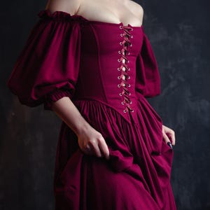 May include: A burgundy off-the-shoulder dress with a fitted bodice and a full skirt. The dress features a lace-up front and puffed sleeves. The model is wearing a black choker necklace. The dress is made of a soft, flowing fabric.