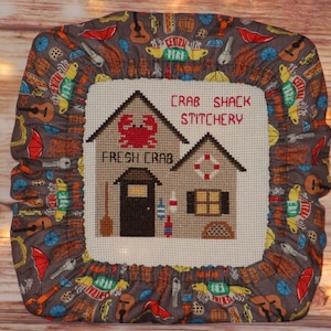 May include: A cross-stitch design featuring a crab shack with the text "FRESH CRAB" and "CRAB SHACK STITCHERY". The design is framed by a gray fabric with a pattern of musical instruments, umbrellas, and other nautical elements.