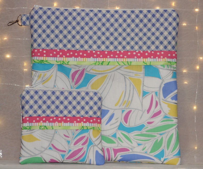 Puede incluir: Un conjunto de dos bolsas de tela. La bolsa m&aacute;s grande tiene una cremallera y un peque&ntilde;o cierre plateado. Ambas bolsas presentan un patr&oacute;n de cuadros azules y blancos, cinta rosa de lunares y un dise&ntilde;o floral con hojas blancas, azules, amarillas y rosas.