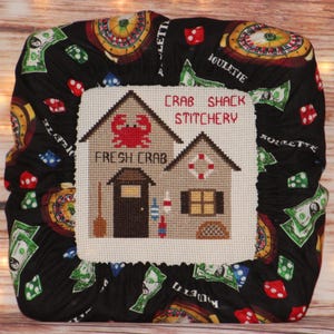 May include: A cross-stitch design featuring a crab shack with the text "CRAB SHACK STITCHERY" and "FRESH CRAB". The design is surrounded by a black fabric patterned with roulette wheels, dice, and dollar bills.