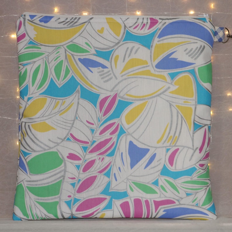 Puede incluir: Bolsa cuadrada con cremallera y un colorido estampado floral. El dise&ntilde;o presenta grandes hojas blancas con contornos grises, acentuadas por elementos amarillos, azules, verdes y rosas sobre un fondo turquesa. Un peque&ntilde;o lazo azul y blanco est&aacute; unido a la cremallera.