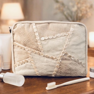 May include: A beige cosmetic bag with a patchwork design, decorated with small, round buttons. Beauty products, including a perfume bottle, lotion tubes, and a toothbrush, are arranged on a wooden surface near the bag.