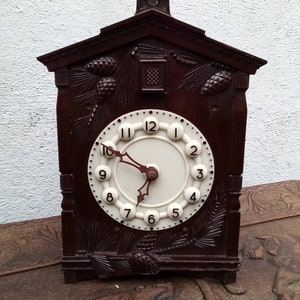 May include: A brown wooden cuckoo clock with a white face and black hands. The clock is decorated with pine cones and branches.