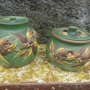 May include: Two green ceramic jars with lids. The jars are decorated with a relief design of leaves, berries, and a small face. The jars are sitting on a yellow and white patterned surface.