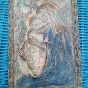 Può includere: Una scultura in ceramica a rilievo incorniciata che rappresenta la Vergine Maria che tiene in braccio un bambino Gesù. La scultura è dipinta in tonalità di blu, marrone e bianco, con accenti dorati. La cornice è in legno e ha un design semplice.