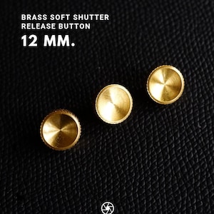 May include: Three brass soft shutter release buttons, each 12 mm in diameter, are displayed on a textured black surface. The buttons have a circular design with a ridged edge and a central, slightly raised area. The text "BRASS SOFT SHUTTER RELEASE BUTTON" and "12 MM." is visible.
