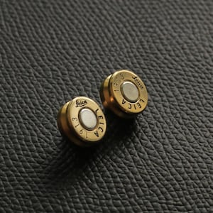May include: Two gold-colored round buttons with the text "Leica 1913" and "Leica" on them. The buttons are on a black textured surface.