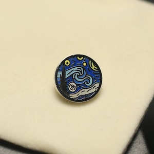 May include: A round, silver-toned pin featuring a miniature depiction of Van Gogh's "Starry Night" painting. The design includes a dark blue sky with swirling yellow stars and a stylized village scene. The pin is set against a neutral background.