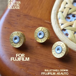 Może przedstawiać: Trzy mosiężne przyciski spustu migawki do aparatów Fujifilm. Każdy przycisk ma srebrny środek i tekst "45 AUTO" oraz "FUJIFILM" wygrawerowany na powierzchni. Widoczny jest również tekst "SOFT SHUTTER BUTTON RELEASE" i "FOR FUJIFILM".
