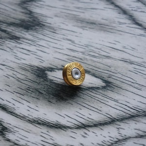 May include: A small, circular, gold-coloured metal object resembling the base of a cartridge casing. The surface features the embossed text FUJIFILM and 45 AUTO, with a central silver-toned circular indentation. The object has a metallic finish.