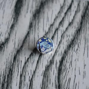 Peut inclure: Une broche émaillée circulaire représentant un ciel nocturne stylisé avec des motifs bleus tourbillonnants, des étoiles jaunes et la silhouette sombre d'un cyprès, le tout entouré d'un bord en métal argenté poli.