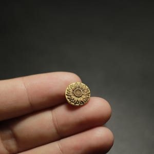 Może przedstawiać: Mały, okrągły, drewniany guzik z szczegółowym wzorem słonecznika wyrzeźbionym na jego powierzchni. Guzik jest w jasnobeżowym kolorze i ma ciemnobrązowy wzór słonecznika. Guzik jest trzymany między dwoma palcami.