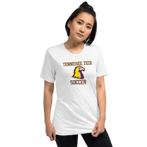 May include: White t-shirt with the text "Tennessee Tech Soccer" in a maroon font. The shirt features a yellow and purple eagle logo. The model is wearing black pants.