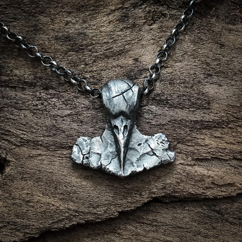 May include: A dark silver-toned pendant shaped like a stylised bird skull with a cracked, weathered texture, suspended from a matching round link chain. The metal surface features deep grooves and a distressed finish throughout the entire piece.