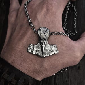 May include: A silver-toned metal pendant shaped like a stylized bird skull or hammer, featuring a textured, weathered surface with deep cracks and crevices. The pendant is suspended from a thick, round-link silver-toned metal chain necklace.