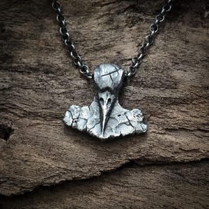 May include: A dark silver-toned metal pendant shaped like a stylized bird skull with a cracked, weathered texture. The pendant is suspended from a matching dark silver-toned metal chain with round links, featuring a rugged, uneven surface finish.