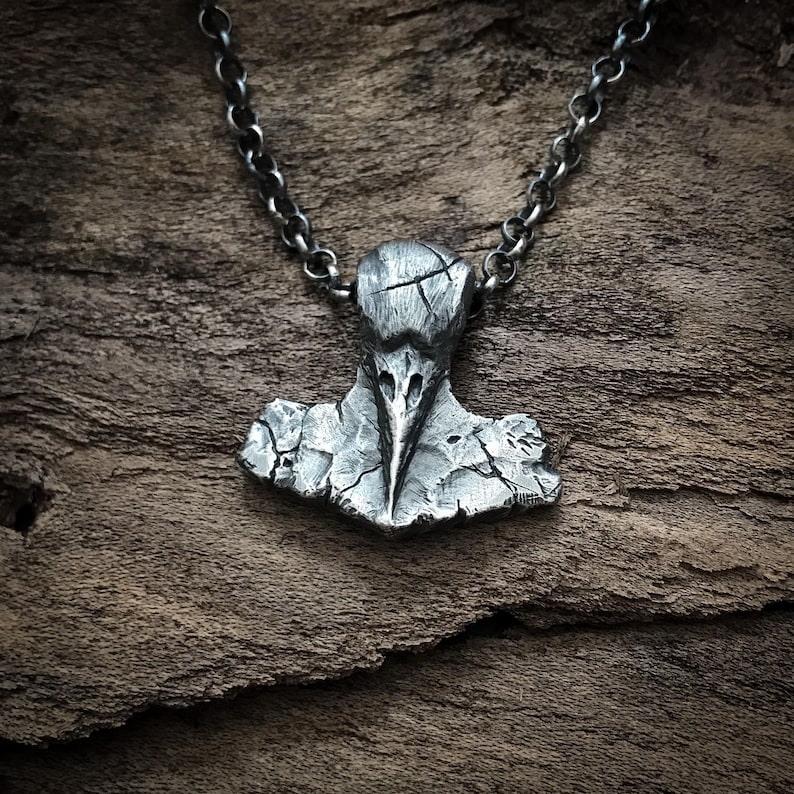 May include: A dark silver-toned metal pendant shaped like a stylized bird skull with a cracked, weathered texture. The pendant is suspended from a matching dark silver-toned metal chain with round links, featuring a rugged, uneven surface finish.