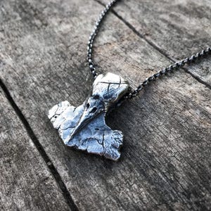 May include: A silver-toned pendant shaped like a bird skull integrated into a hammer-like form, suspended from a dark metal chain. The pendant features a highly textured, weathered surface with deep crevices and a metallic, oxidized finish.
