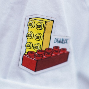 May include: White t-shirt with a red and yellow embroidered patch of a LEGO brick tower with the word "DENMARK" on it.